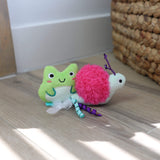 Two plush Quirky Kitty Glow Critters— a green frog and a pink snail—sit on a wooden floor by a basket. Filled with catnip and silvervine, these glow-in-the-dark toys are perfect for playful cats. Available in a convenient 2-pack.