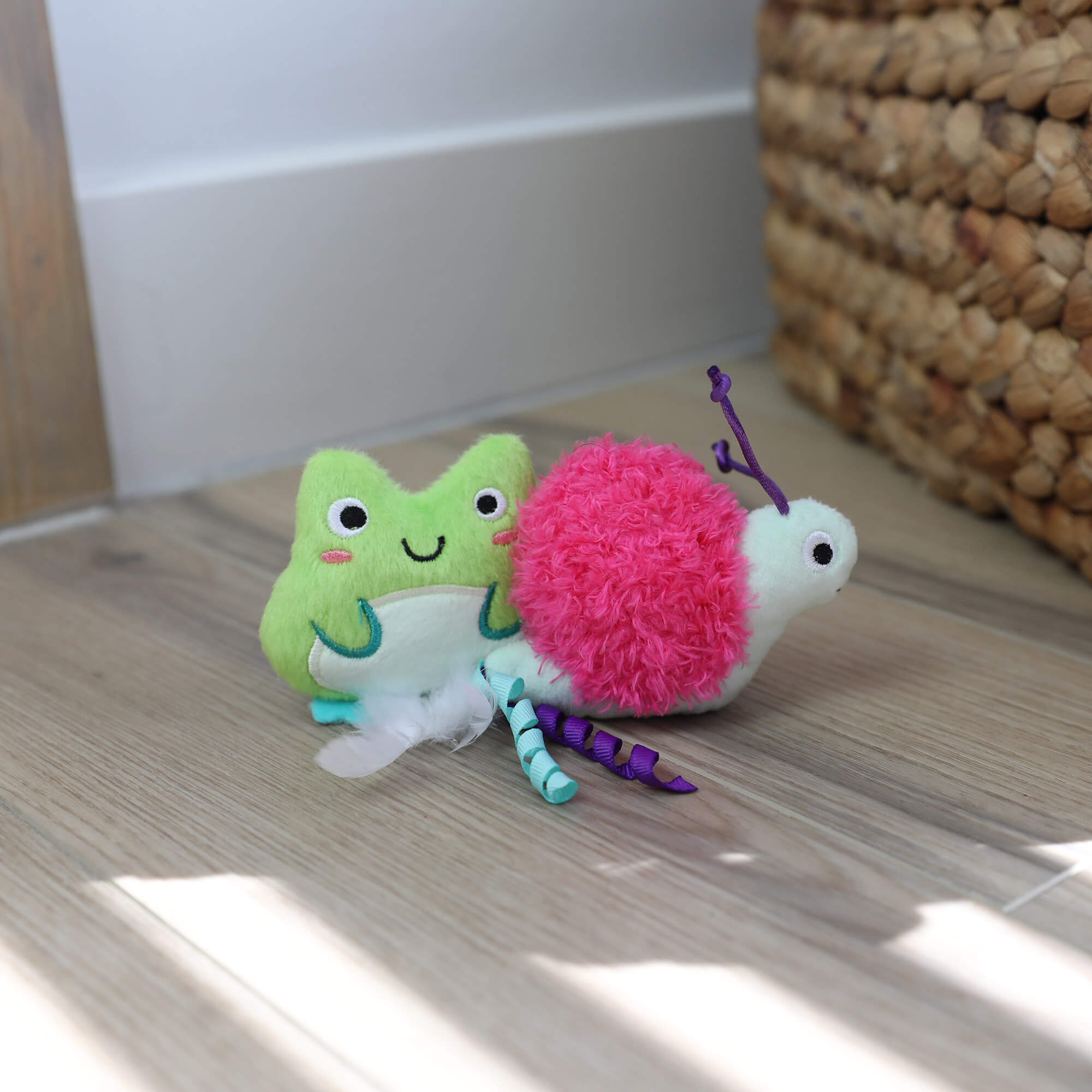 Two plush Quirky Kitty Glow Critters— a green frog and a pink snail—sit on a wooden floor by a basket. Filled with catnip and silvervine, these glow-in-the-dark toys are perfect for playful cats. Available in a convenient 2-pack.