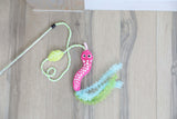 The Quirky Kitty Glow Snake Wand – Glowing Forest Cat Toy with Catnip & Silvervine by Quirky Kitty, featuring googly eyes, ruffled ribbons, and a green leaf, lies on a light wood floor.