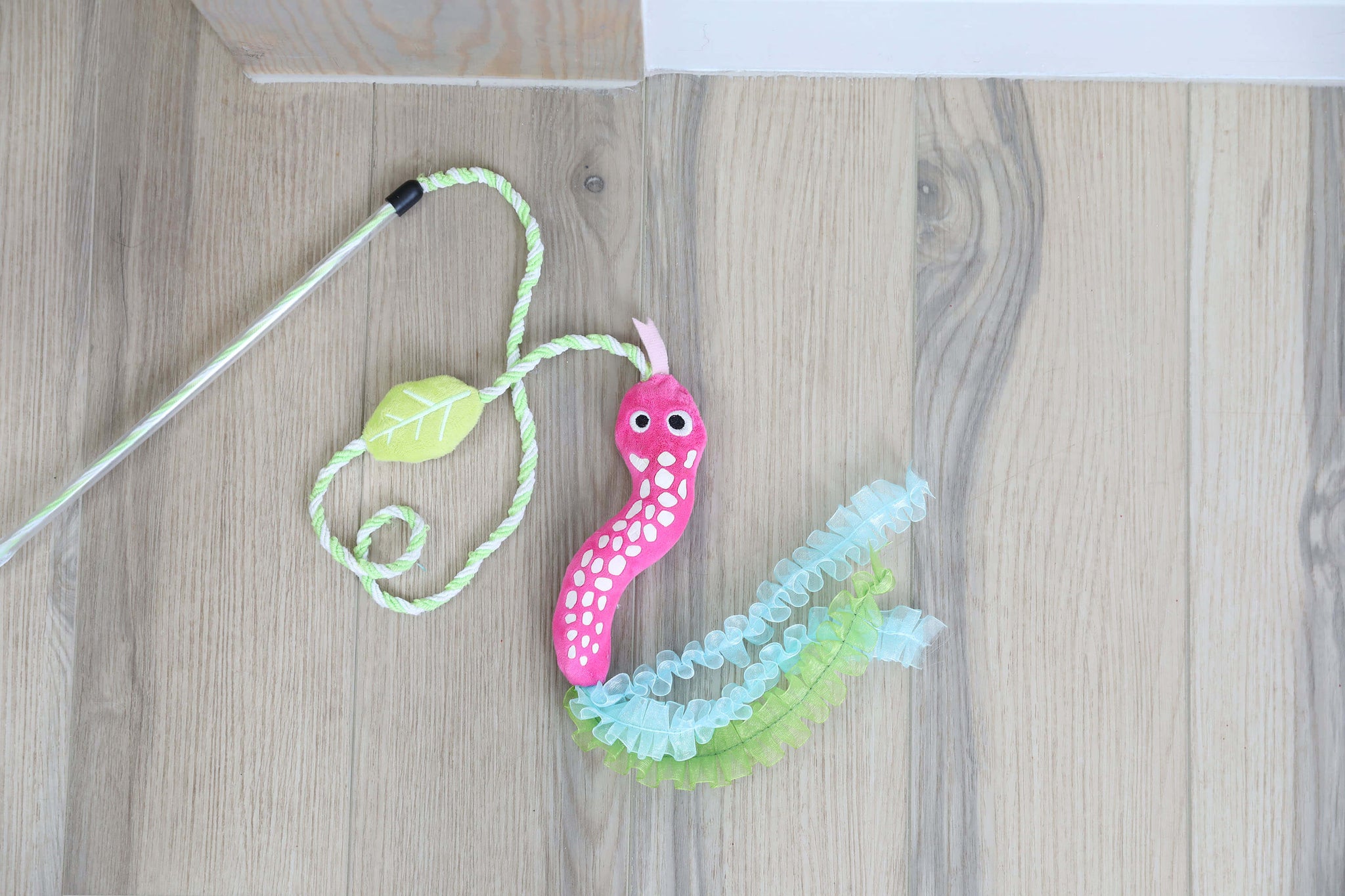The Quirky Kitty Glow Snake Wand – Glowing Forest Cat Toy with Catnip & Silvervine by Quirky Kitty, featuring googly eyes, ruffled ribbons, and a green leaf, lies on a light wood floor.