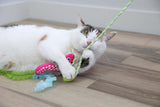 A white and tabby cat lies on a wooden floor, hugging the Quirky Kitty Glow Snake Wand by Quirky Kitty, a glowing forest cat toy with catnip and silvervine, while playing contentedly with its green striped string.