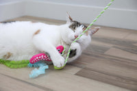 A white and tabby cat lies on a wooden floor, hugging the Quirky Kitty Glow Snake Wand by Quirky Kitty, a glowing forest cat toy with catnip and silvervine, while playing contentedly with its green striped string.