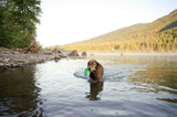In a tranquil lake, a brown dog holds a Chuckit! Amphibious Boomerang by Chuckit in its mouth. Mist rises from the water against a backdrop of rocky shores and tree-covered hills beneath a clear sky.