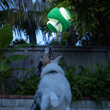 A fluffy-coated dog jumps excitedly in a garden with mouth open, aiming for the Chuckit! Kick Fetch Max Glow. This interactive toy from Chuckit glows green and white in mid-air against a backdrop of lush greenery and a wooden fence.