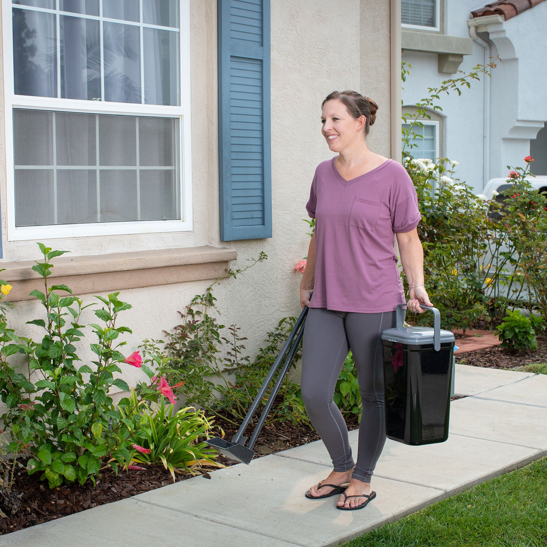 A person in a mauve shirt and gray leggings is smiling on their lawn clean-up routine, holding the PetFusion Portable Outdoor Pet Waste Disposal System. They are on a sidewalk beside a house with flower-adorned bushes under windows with blue shutters.