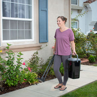 A person in a mauve shirt and gray leggings is smiling on their lawn clean-up routine, holding the PetFusion Portable Outdoor Pet Waste Disposal System. They are on a sidewalk beside a house with flower-adorned bushes under windows with blue shutters.