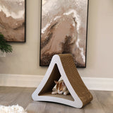 A cat sleeps in the PetFusion 3-Sided Vertical Cat Scratching Post, featuring a recycled cardboard triangular design. Its sleek white frame sits on a wooden floor. Behind it, abstract art with brown and white swirls decorates the wall, while a plant and fluffy rug are partly visible nearby.