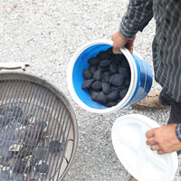 A person stands beside a blue Vittles Vault filled with charcoal, its gravel contrast highlighted by beige shoes and a plaid shirt. The Vittles Vault 2.0 Replacement Lid is nearby as they prepare to grill, coals already heating up.