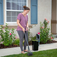 A woman in a purple shirt and gray leggings uses the PetFusion Portable Outdoor Pet Waste Disposal System on the lawn. Nearby, a portable dog waste station stands ready. The setting includes a beige house with blue shutters and flowering bushes.