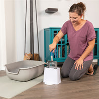 Wearing a purple shirt, someone smiles while using the PetFusion 2-in-1 Universal Litter Scoop Holder on a waste container beside a cat litter box. A curious cat watches nearby, with a teal cabinet and lamp adding charm to the scene.