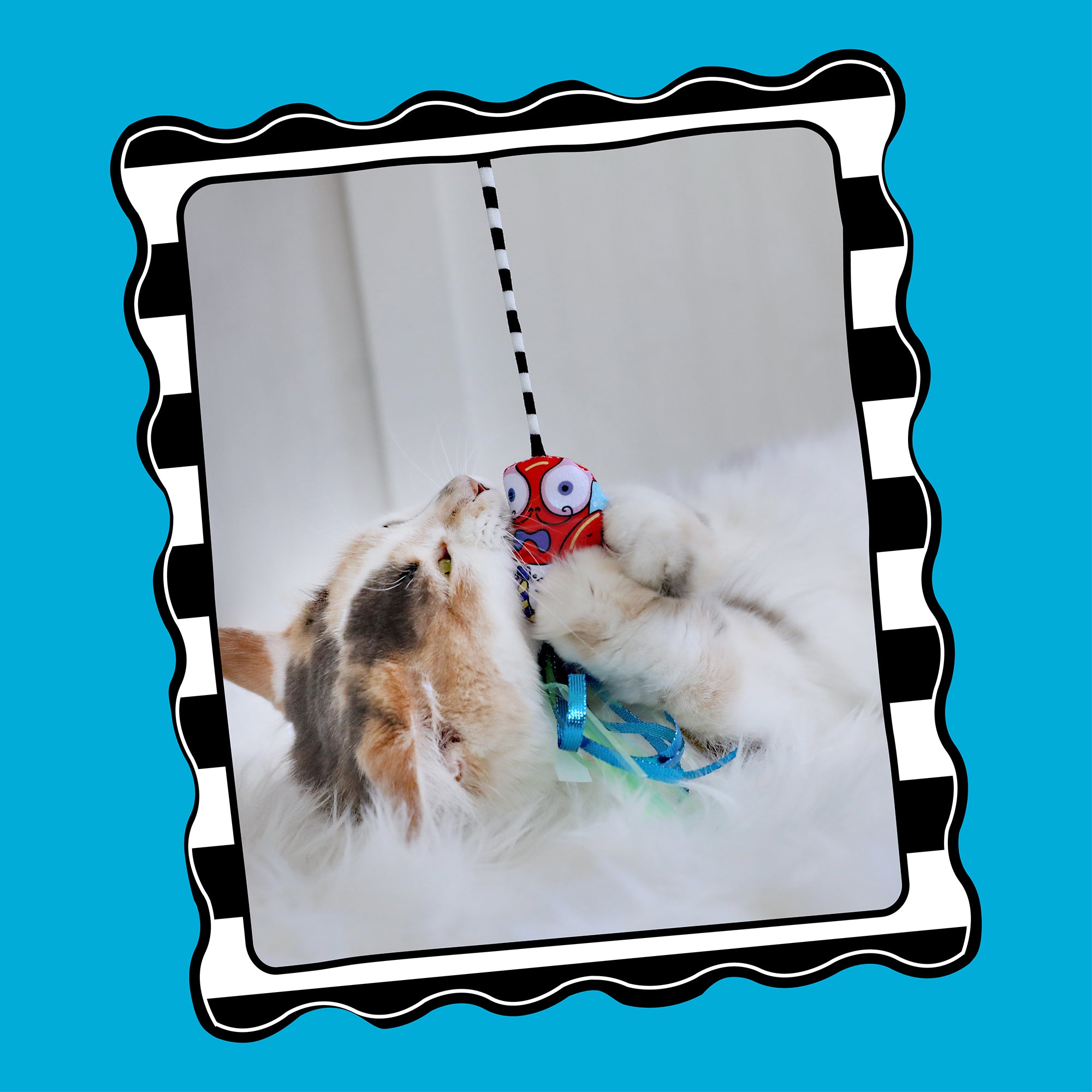 A cat playfully interacts with a red toy from the FAT CAT Catfisher Teasers - Crawdaddy Wand by Fatcat, hanging from a black and white striped string above a fluffy white blanket. The scene is framed by a wavy black and white border on a blue background.