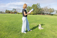 A woman stands on a grassy field holding the Chuckit! Cannon & Cannon Max Dog Ball Launcher by Chuckit!, as a small white and tan dog eagerly sits in front of her. Trees and blue sky appear in the background.