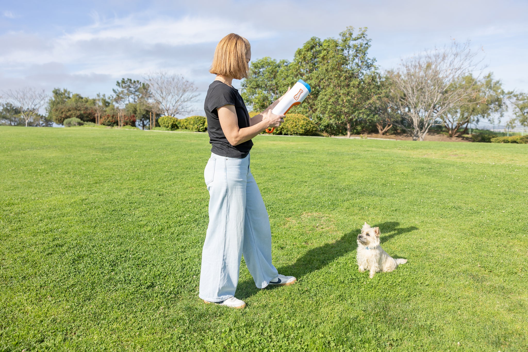 A woman stands on a grassy field holding the Chuckit! Cannon & Cannon Max Dog Ball Launcher by Chuckit!, as a small white and tan dog eagerly sits in front of her. Trees and blue sky appear in the background.