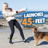 A woman on a beach uses the Chuckit! Cannon & Cannon Max - Dog Ball Launcher from Chuckit! as her dog watches. Large text reads, Launches 45+ feet. The ocean and rocks are in the background.
