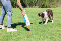 Wearing jeans and white sneakers, a person uses the Chuckit! Cannon & Cannon Max Dog Ball Launcher by Chuckit! to play fetch with a brown and white dog on a grassy field, as the dog eagerly watches the ball. Trees and a fence are in the background.