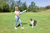 A woman in sunglasses stands on a grassy field aiming the Chuckit! Cannon & Cannon Max Dog Ball Launcher by Chuckit! as her brown and white dog looks up eagerly, ready for an exciting fetch session with their favorite toy under a clear sky.