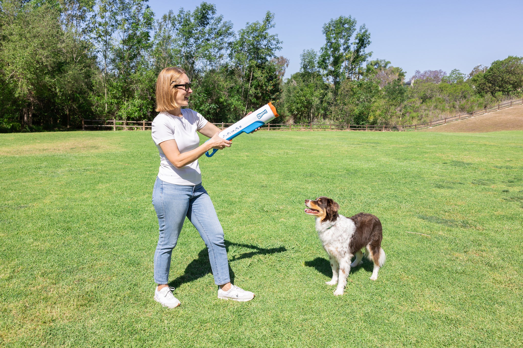 A woman in sunglasses stands on a grassy field aiming the Chuckit! Cannon & Cannon Max Dog Ball Launcher by Chuckit! as her brown and white dog looks up eagerly, ready for an exciting fetch session with their favorite toy under a clear sky.