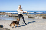 A woman in a white sweater and black leggings stands on a sandy beach holding the Chuckit! Cannon & Cannon Max Dog Ball Launcher by Chuckit!, as a brown and white dog looks up at her with the ocean and rocks in the background under a clear blue sky.