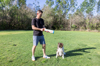 A man in a black T-shirt and shorts stands on grass holding the Chuckit! Cannon & Cannon Max Dog Ball Launcher by Chuckit!, while a brown and white dog sits nearby, looking up. Trees and a wooden fence are visible in the background.