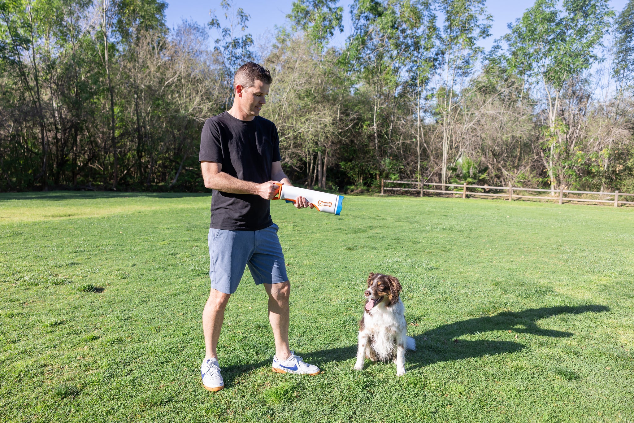 A man in a black T-shirt and shorts stands on grass holding the Chuckit! Cannon & Cannon Max Dog Ball Launcher by Chuckit!, while a brown and white dog sits nearby, looking up. Trees and a wooden fence are visible in the background.