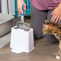 A person in leggings uses the PetFusion 2-in-1 Universal Litter Scoop Holder with durable silicone bristles, as a cat sniffs nearby. This scene unfolds on a wooden floor next to a gray litter mat and litter box.