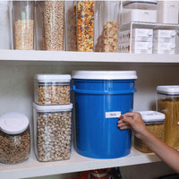 A pantry shelf neatly displays containers of cereals, pasta, and nuts. A prominent large blue Vittles Vault 2.0 Replacement Lid labeled Rice stands out as a person holds a label beside it.