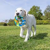 A white dog joyfully carries the Chuckit! EcoFetch Recycled Rubber Flyer by Chuckit in its mouth while walking on a lush green lawn, with trees and a clear blue sky in the background.