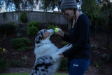 A woman in a beanie and glasses playfully holds a Chuckit Max Glow Ultra Squeaker Ball as she interacts with a black-and-white-coated standing dog in a garden, surrounded by lush trees and plants.