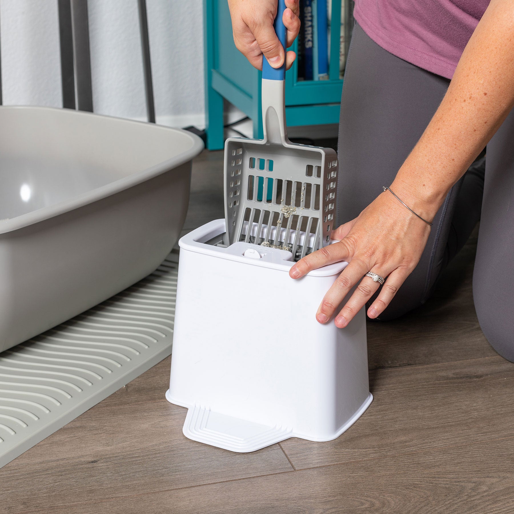 Kneeling on a wooden floor, a person in a pink shirt and gray pants efficiently sifts litter in a white container next to a gray box using the blue-handled PetFusion 2-in-1 Universal Litter Scoop Holder, which has durable waterproof hypoallergenic silicone bristles.