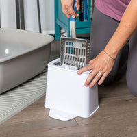 Kneeling on a wooden floor, a person in a pink shirt and gray pants efficiently sifts litter in a white container next to a gray box using the blue-handled PetFusion 2-in-1 Universal Litter Scoop Holder, which has durable waterproof hypoallergenic silicone bristles.