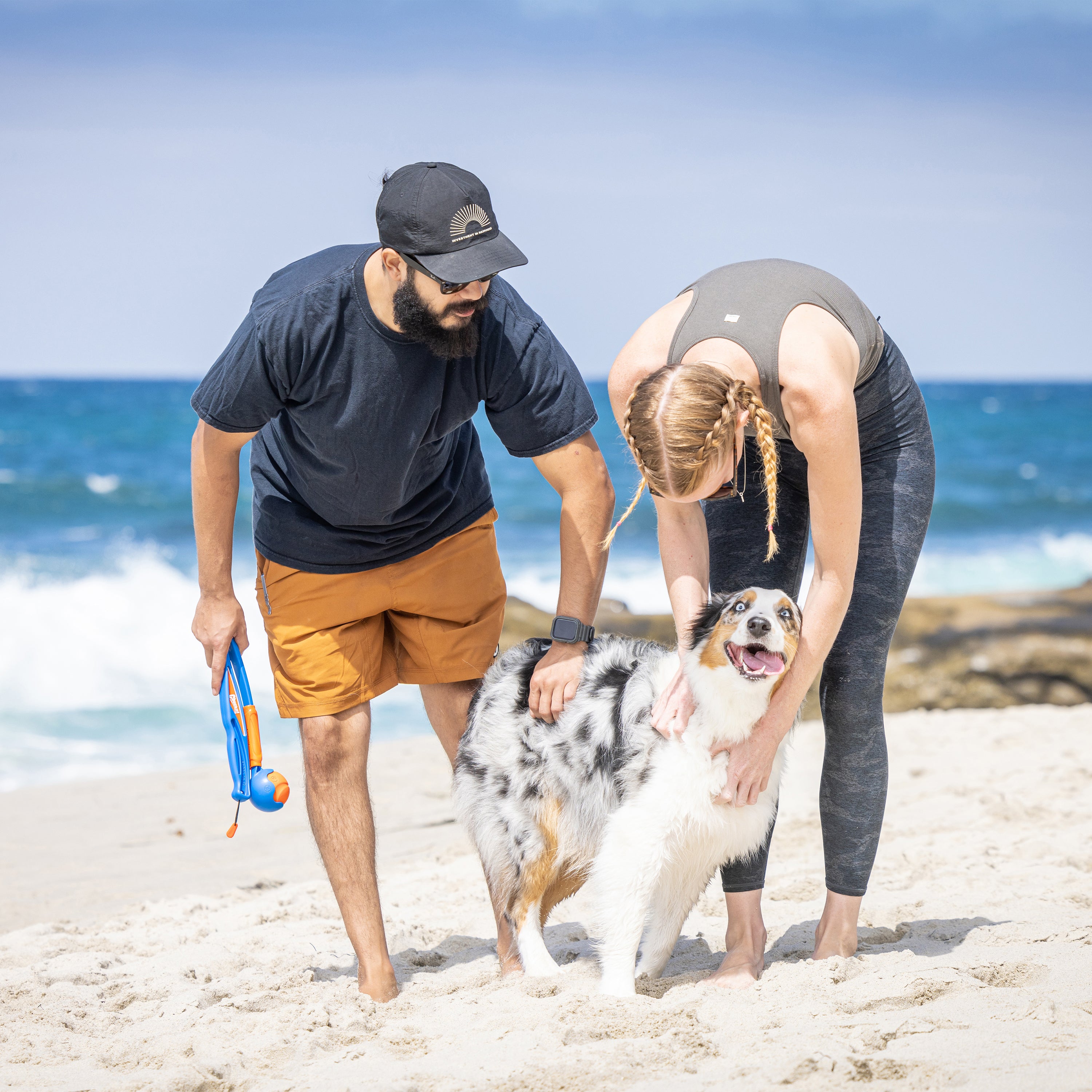A couple plays joyfully with their happy dog on a sandy beach. The man uses the Chuckit Fetch & Fold Launcher from Chuckit, while the woman pets the dog. Ocean waves dance under a clear blue sky, creating their perfect dog park.