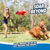Wearing athletic gear, a woman throws a Chuckit! Flying Squirrel fetch toy to her golden retriever, darting across the grass. The text reads SOAR BEYOND and Glides through the air for easy tracking and chasing. Trees and a fence frame this joyful scene of play.