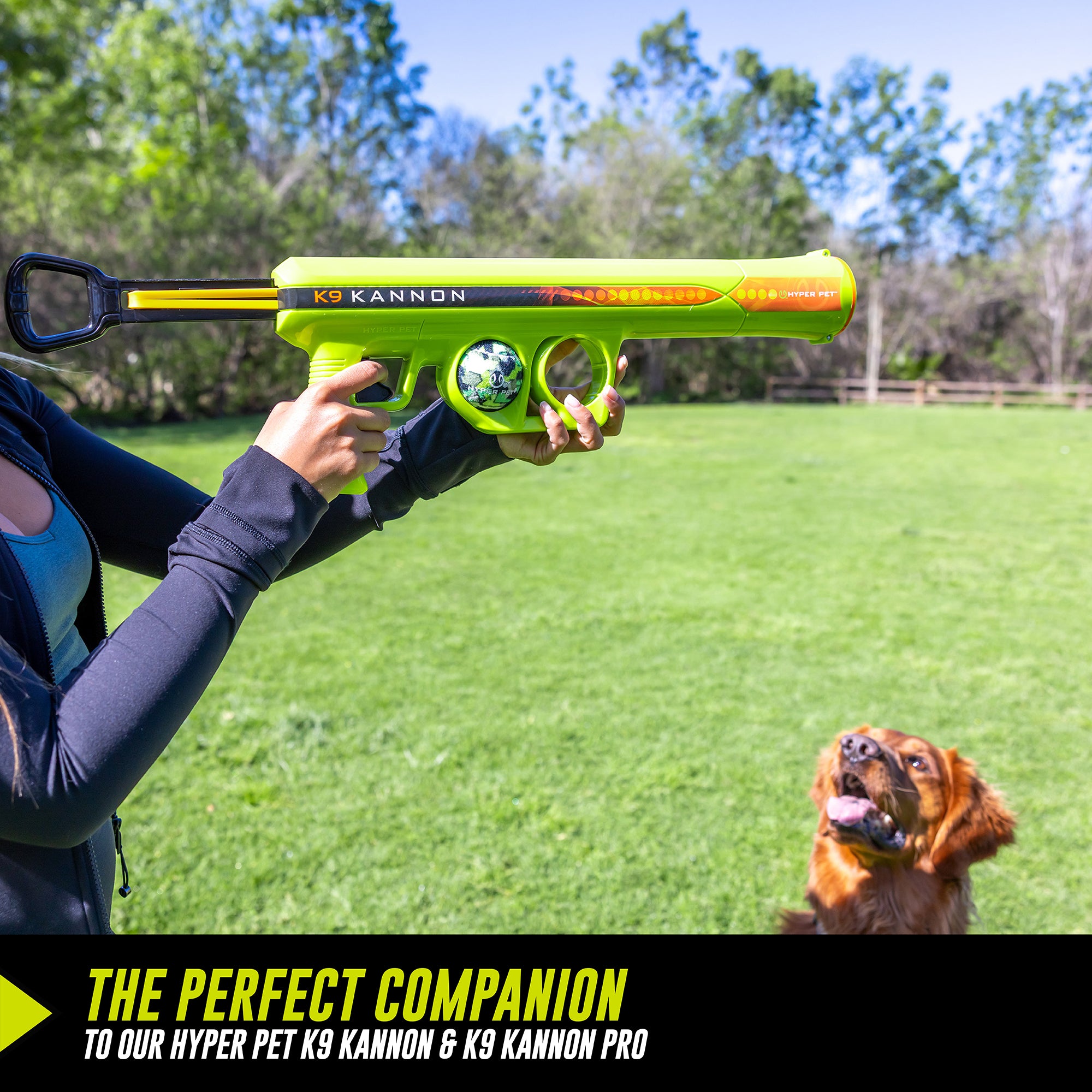 A person outdoors holds a green and orange dog toy launcher, aiming it as a brown dog eagerly waits in the grassy field. Text at the bottom promotes Hyperpets K9 Kannon as the perfect companion for pets. Trees and a wooden fence are in the background.