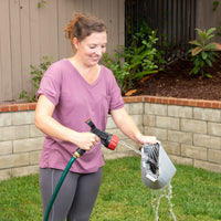 A woman in a purple shirt and gray pants, smiling, effortlessly hoses down a fan on the grass by a plant-covered brick wall. The waterproof hypoallergenic silicone of the PetFusion 2-in-1 Universal Litter Scoop Holder keeps it fresh like her joyful demeanor.
