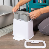 A person assembles the white PetFusion 2-in-1 Universal Litter Scoop Holder with a removable top and handle-like component made from waterproof hypoallergenic silicone. Its positioned on a wooden floor next to a gray litter box and mat, adding practicality and style to the home interior.