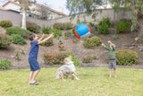 Two boys play with a Chuckit! Big Air Fetch & Herding Ball Dog Toy in a grassy backyard as their dog leaps up excitedly, herding the Chuckit ball. Shrubs and trees line the background.