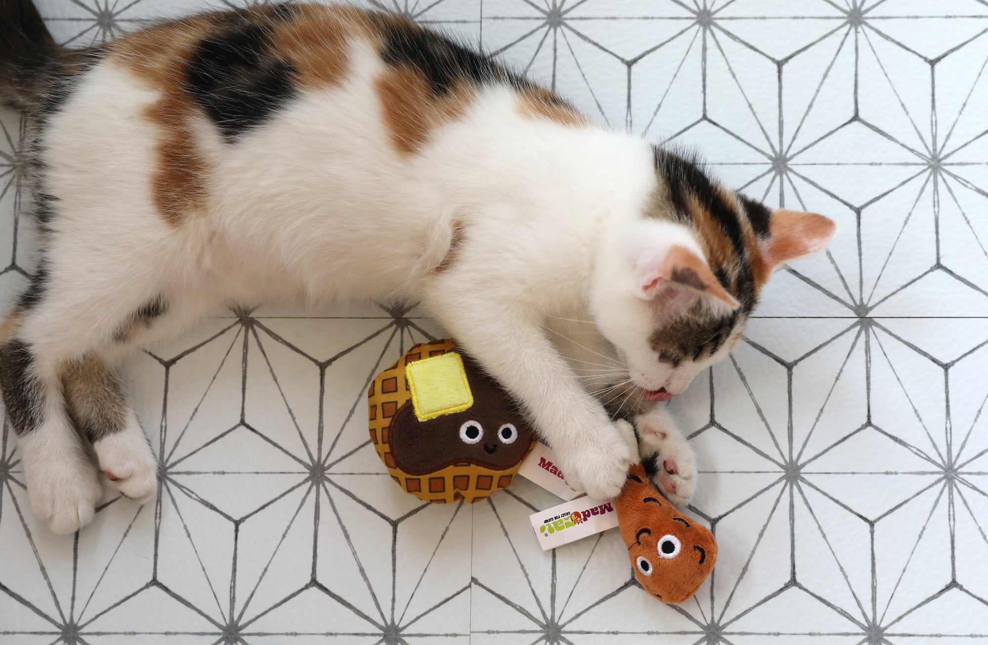 A calico kitten playfully bats at Mad Cats Chicken and Waffles Twin Pack toys, shaped like a waffle and syrup bottle. Both toys have cute cartoon faces and are infused with catnip and silvervine for extra fun on a geometric-patterned floor.