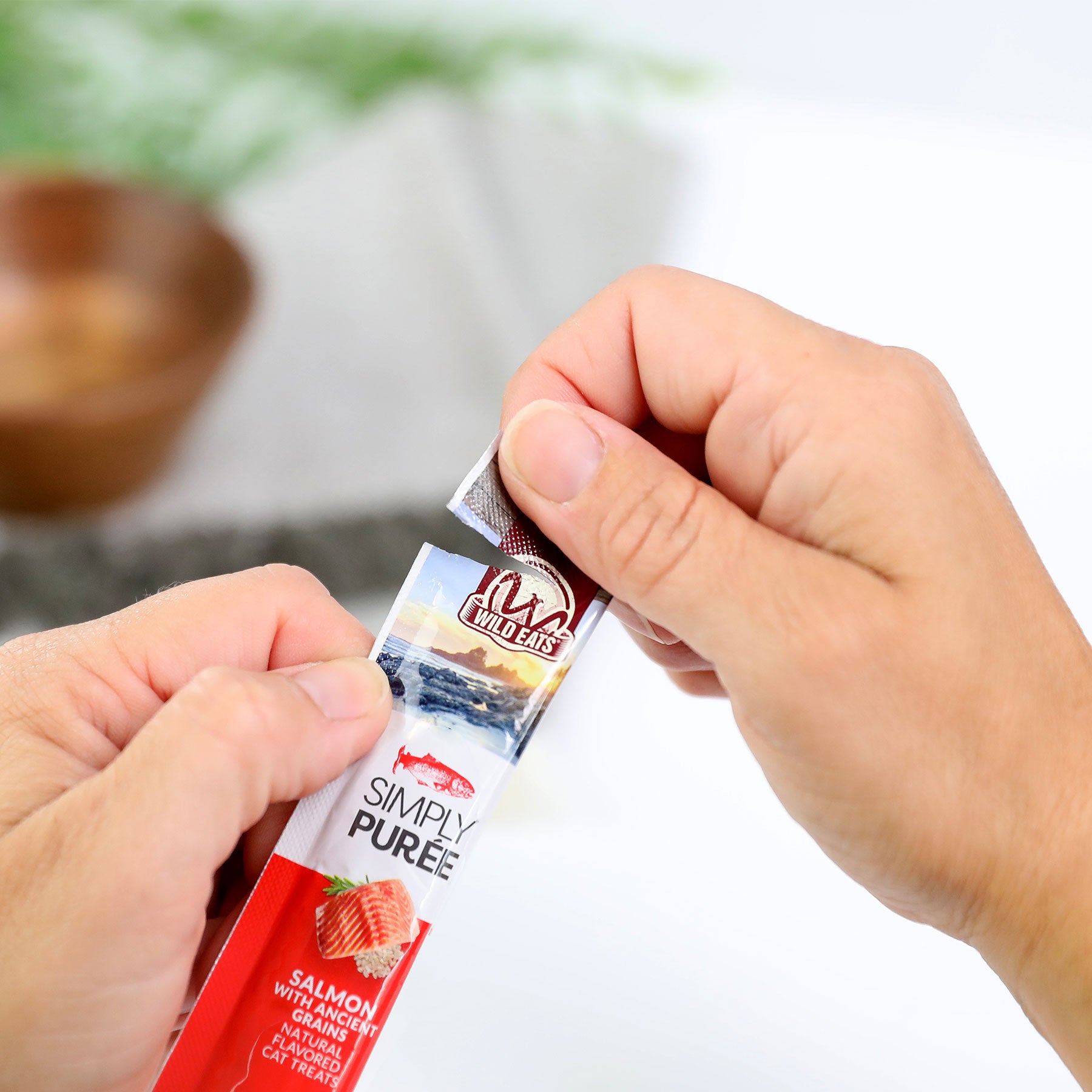 Opening a red and white Wild Eats Simply Puree Salmon Cat Treat packet reveals Omega-3 rich treats adorned with a pet face logo alongside a salmon image; a wooden bowl is in the blurred background.