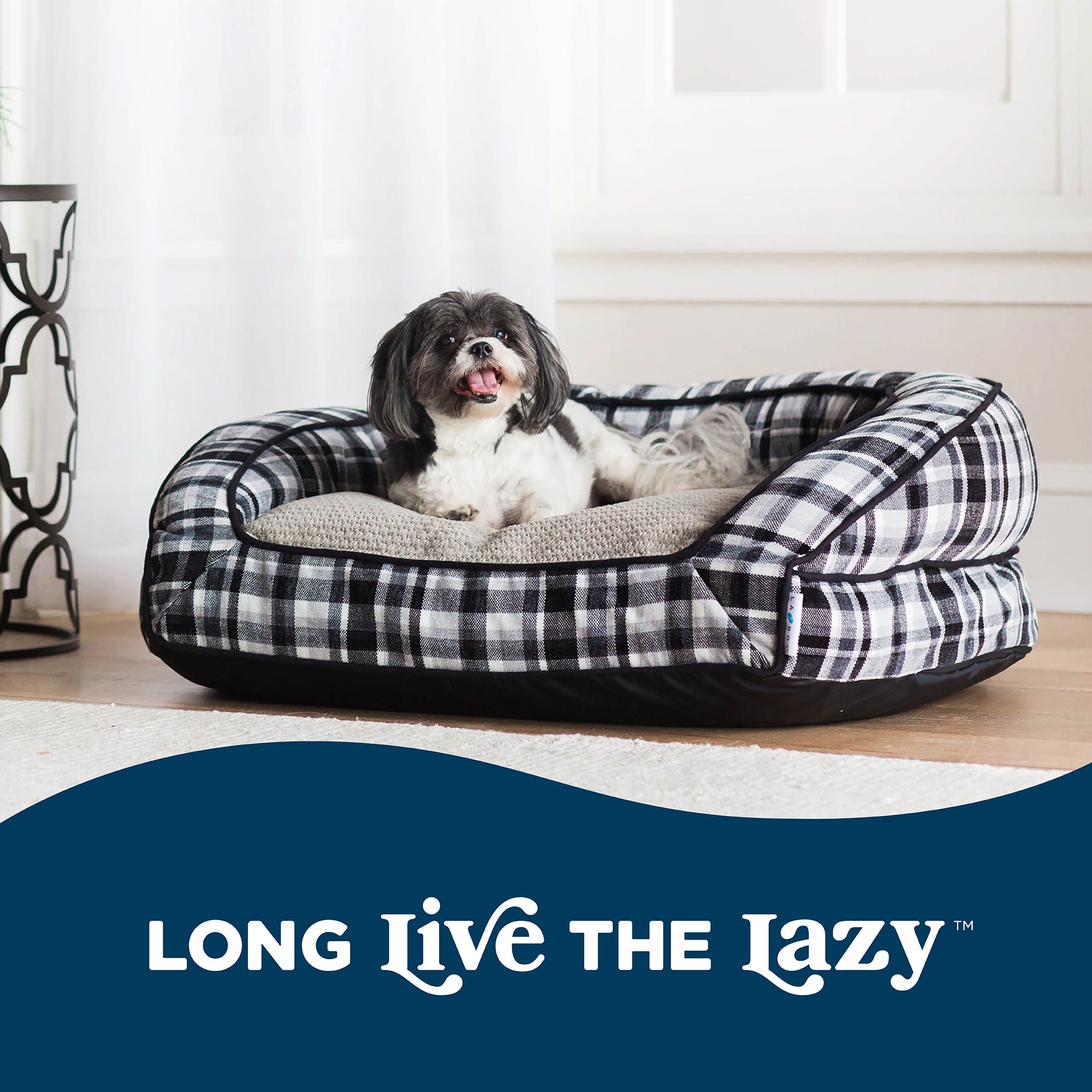 A small black and white dog relaxes on a La-Z-Boy Tucker Plaid Sofa Dog Bed, featuring a ComfortSelect fiberfill cushion. The room is bright with curtains in the background, and text reads Long Live The Lazy™. Ideal for medium-sized dogs!.