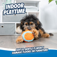 A fluffy dog joyfully plays with a Chuckit! Indoor Ball—orange and blue with Bounceflex Core—on a wooden floor. A white shelf unit is in the background. Text reads INDOOR PLAYTIME and Soft & gentle, it wont damage floors or furniture.