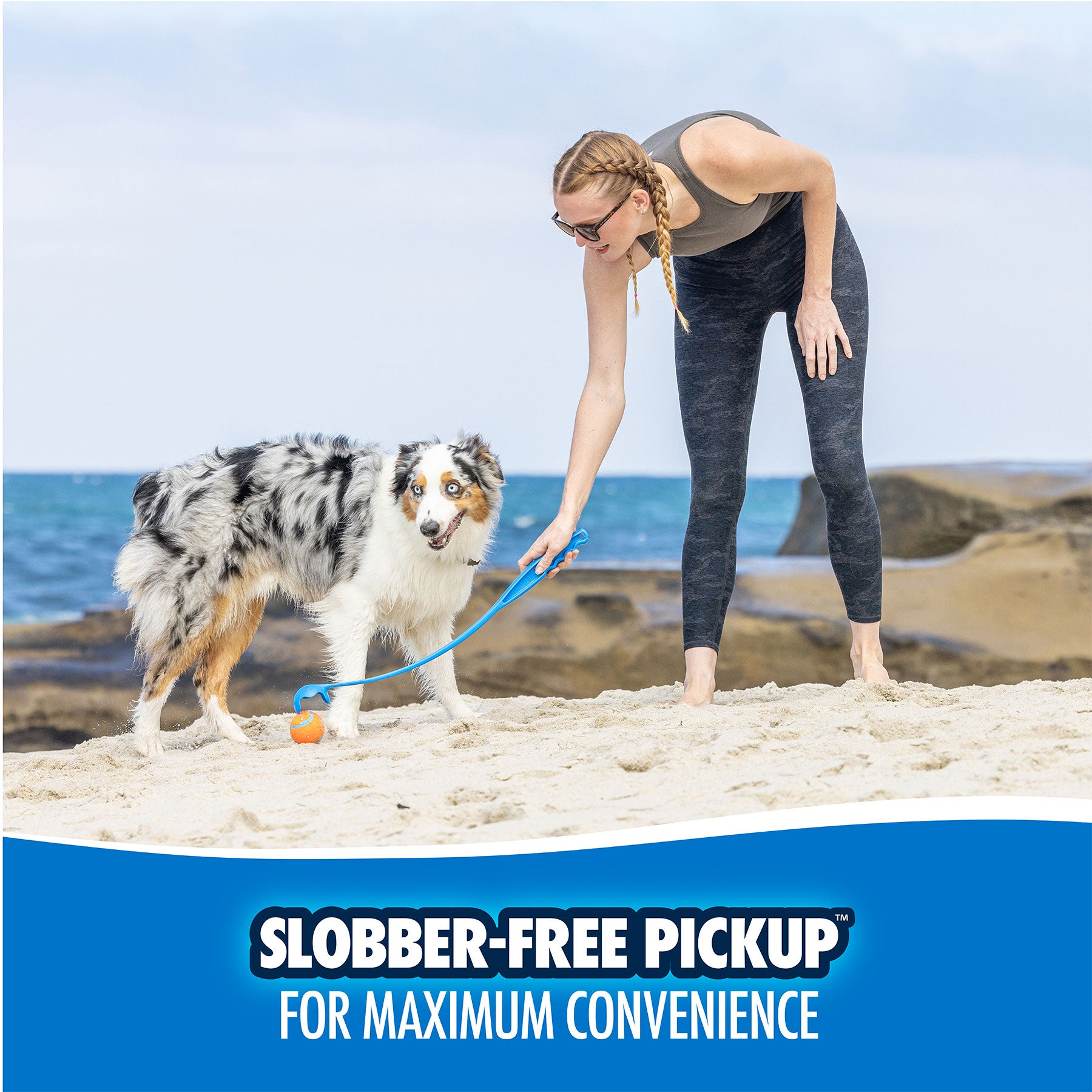 A woman in athletic wear plays fetch with a dog on a sandy beach using the ergonomic Chuckit! Pro Launcher. Ocean and rocks are in the background, with a banner reading Slobber-Free Pickup for Maximum Convenience.