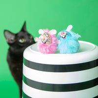 A black cat curiously observes two FAT CAT Classic Fluff Bunnies from Fatcat, one pink and one blue, sitting on a black-and-white striped container infused with organic catnip. This playful kitty is ready to zoom around, set against a solid green background.