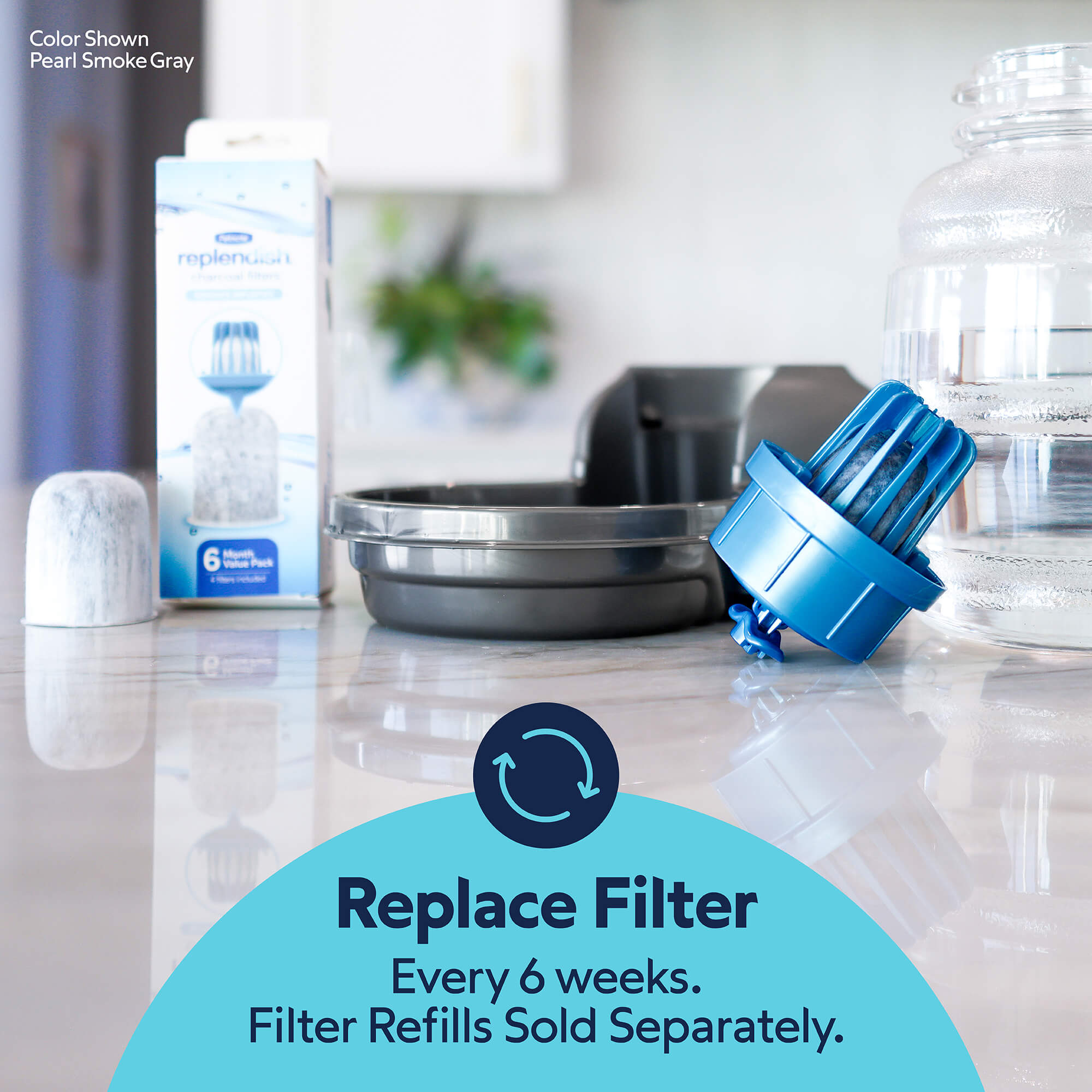 The Petmate Replendish 0.5 Gallon Filtered Pet Waterer with Gravity Refill sits on a counter with a pitcher, filter box, and glass jar. Replace filter every 6 weeks for ideal pet hydration. Filter refills sold separately.