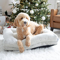 A fluffy dog relaxes on a La-Z-Boy Salt & Pepper Rosie Lounger Dog Bed near a Christmas tree with white ornaments. Wrapped gifts and a cozy brown armchair sit on a plush white rug in the background.