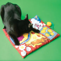 A black cat joyfully plays on a vibrant FAT CAT Big Mamas Makin Biscuits Boogie Mat by Fatcat, tapping its paws on the cartoon prints against a green background. Nearby, a small yellow toy infused with organic catnip is ready to join the fun.