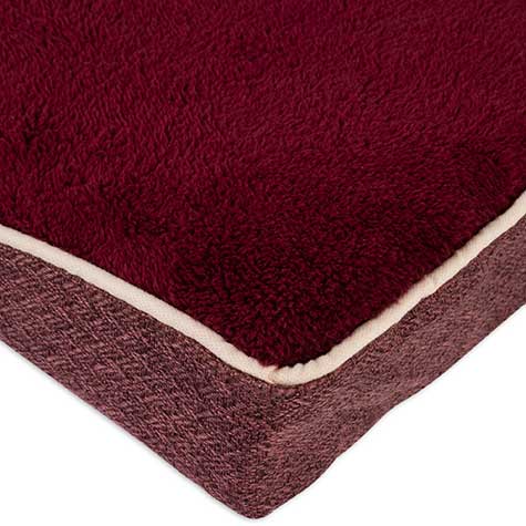 A close-up of the Aspen Pet Burgundy Orthopedic Pet Bed shows a corner with dark red, textured micro-lux fabric. It features beige trim and a supportive base made of darker mattress-like foam.