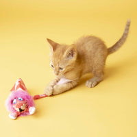 A small orange kitten eagerly plays with a Fatcat FAT CAT Classic Tailchasers Cat Toy against a bright yellow background, reaching out to touch the colorful, fuzzy toy adorned with pink fur and cartoonish eyes.