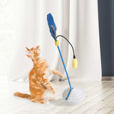 A ginger cat playfully stands on its hind legs reaching for a blue feather teaser toy attached to a flexible stand with a white base near sheer white curtains in a room with light wood flooring, alongside the JW Catnip Mister by Jw.