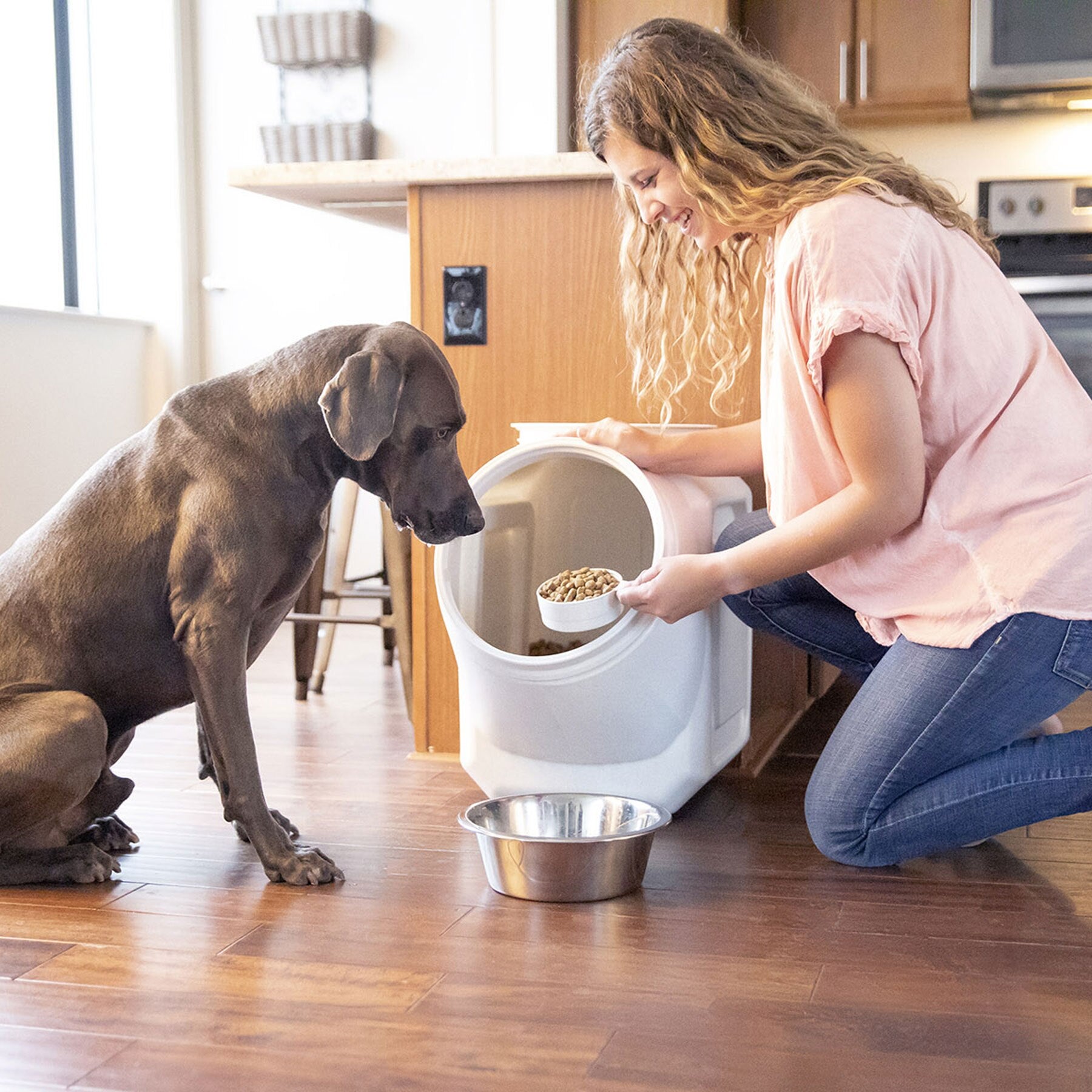 A woman kneels on a wooden floor, scooping dog food from a Vittles Vault Outback Stackable Pet Food Storage Container into a metal bowl. A large brown dog sits attentively, enjoying the freshness provided by the airtight Gamma Seal Technology in this container.