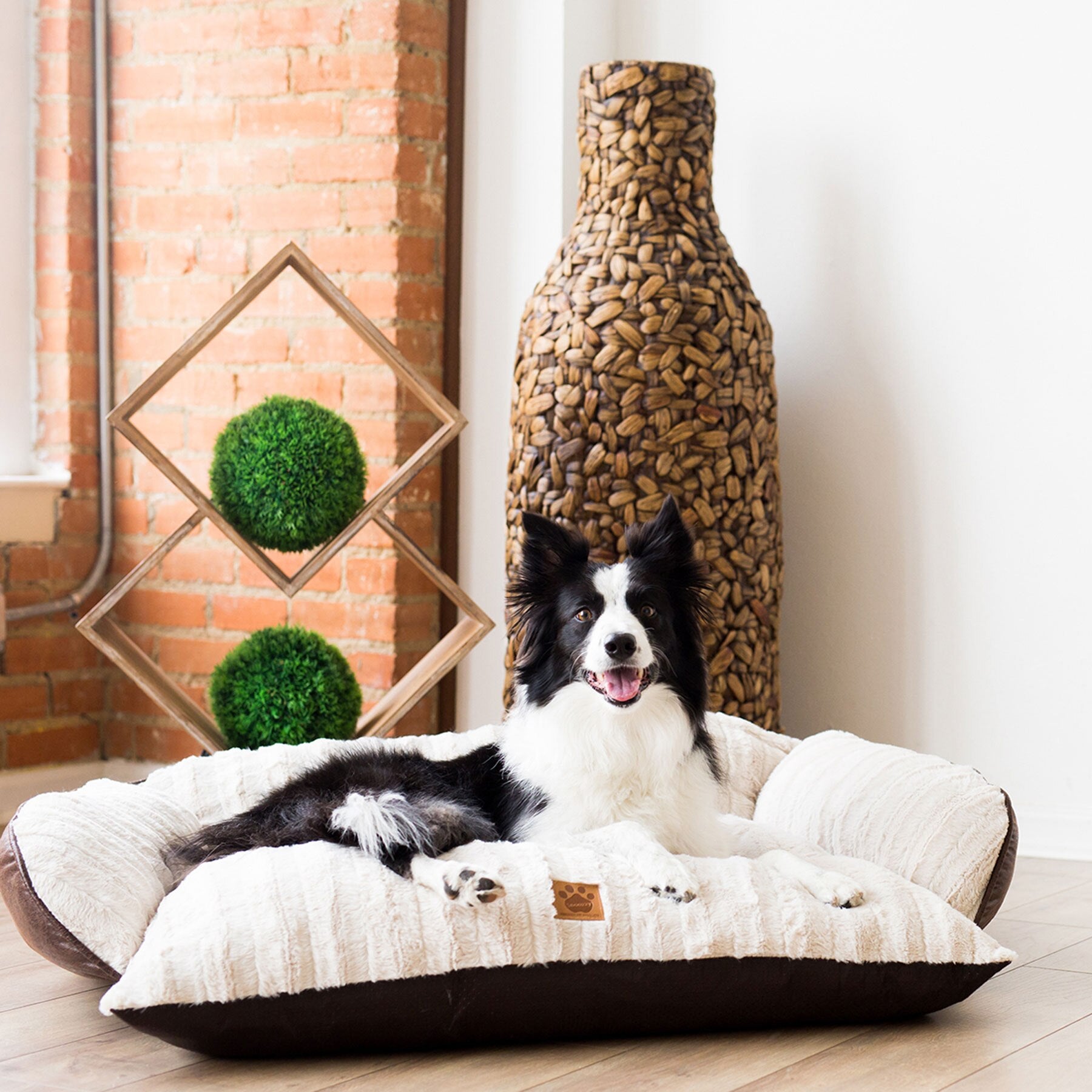 A black and white dog sits happily on a plush white SnooZZy Rustic Luxury Comfy Couch Pet Bed by Snoozzy in a room with a brick wall. A textured vase and geometric wooden sculpture with green spheres enhance the background. The bed is machine washable, blending relaxation with practicality.